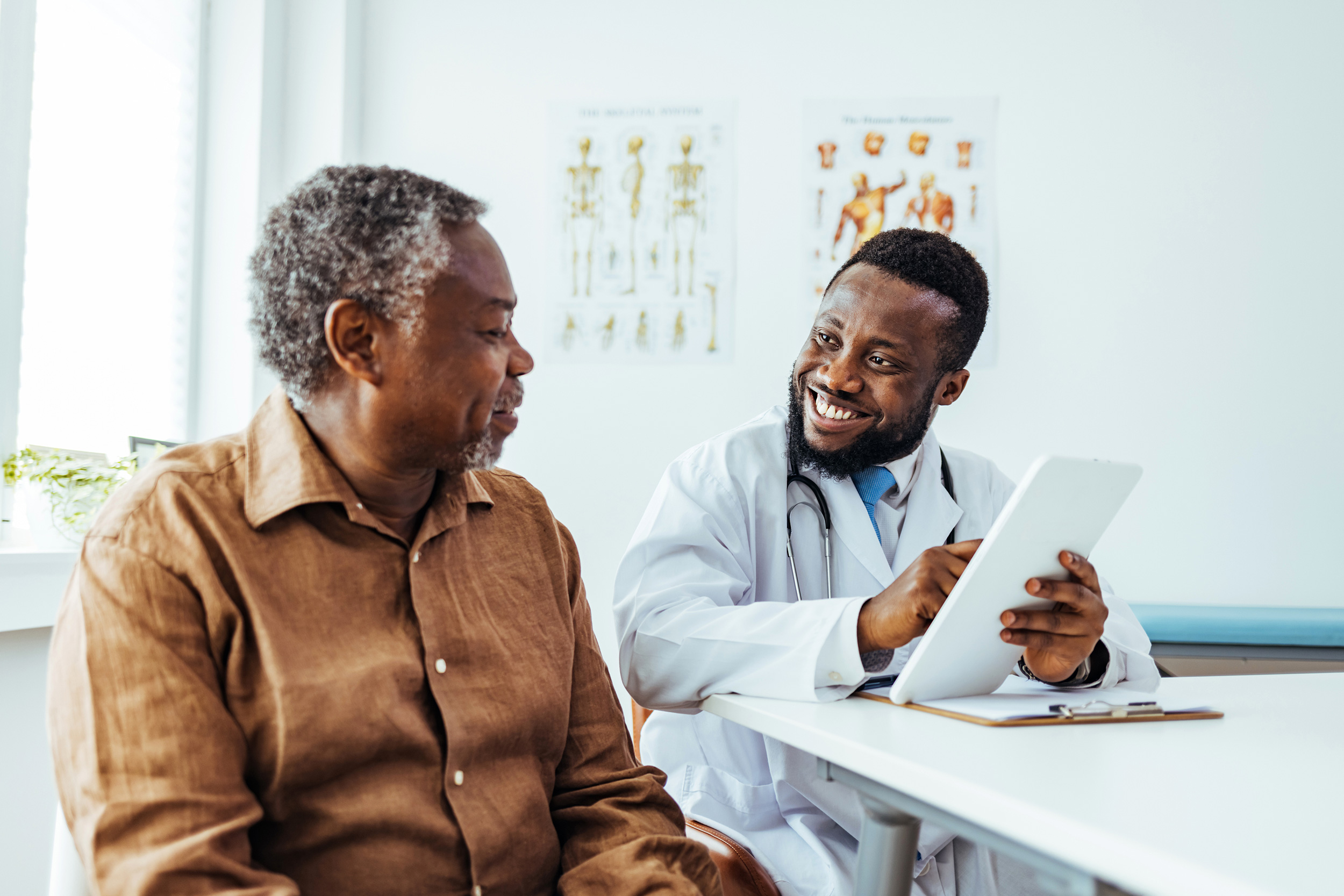 Sitting at a table in a clinic office, a man talks to his doctor, who wears his medical coat and stethoscope while holding a tablet computer. When health problems lead to job loss, Social Security Disability lawyers help people get financial relief.