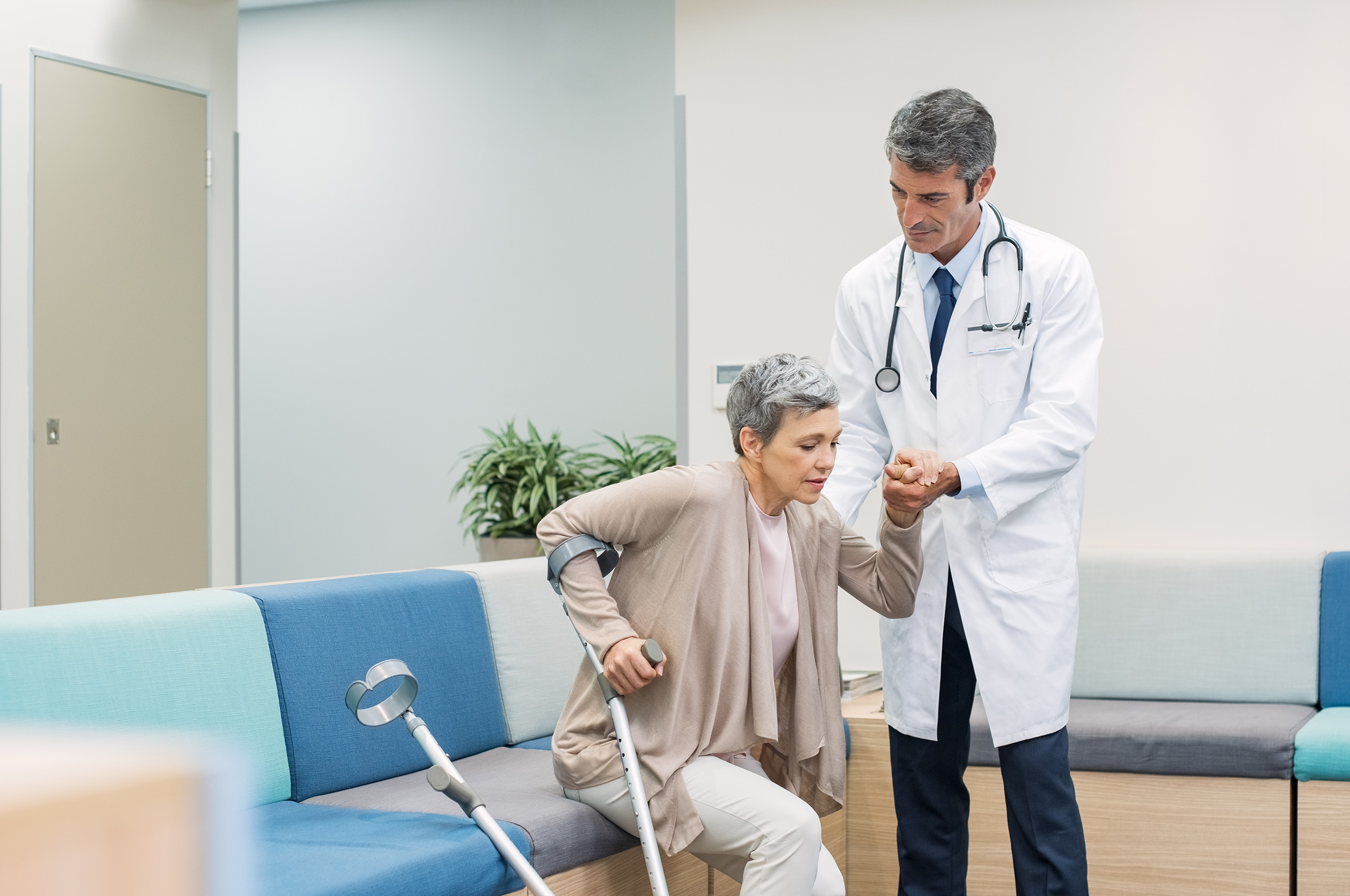 A doctor with a medical coat and stethoscope helps a woman with crutches get up from a seat in a waiting room. Many impairments qualify for Social Security Disability.