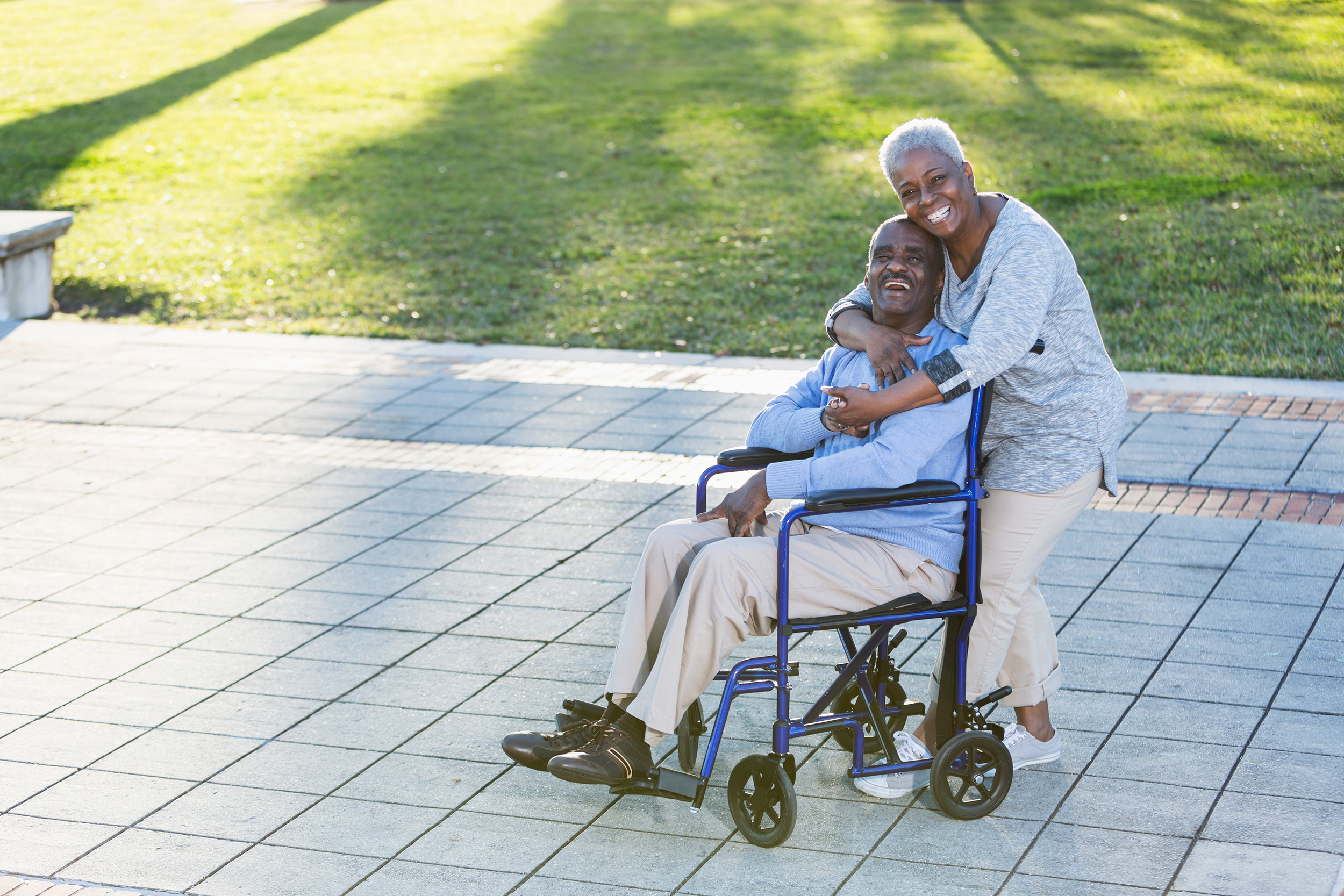 Standing outdoors, a woman hugs a man from behind as he sits in his wheelchair, and they smile. SSI benefits pay monthly amounts set each year by the government.