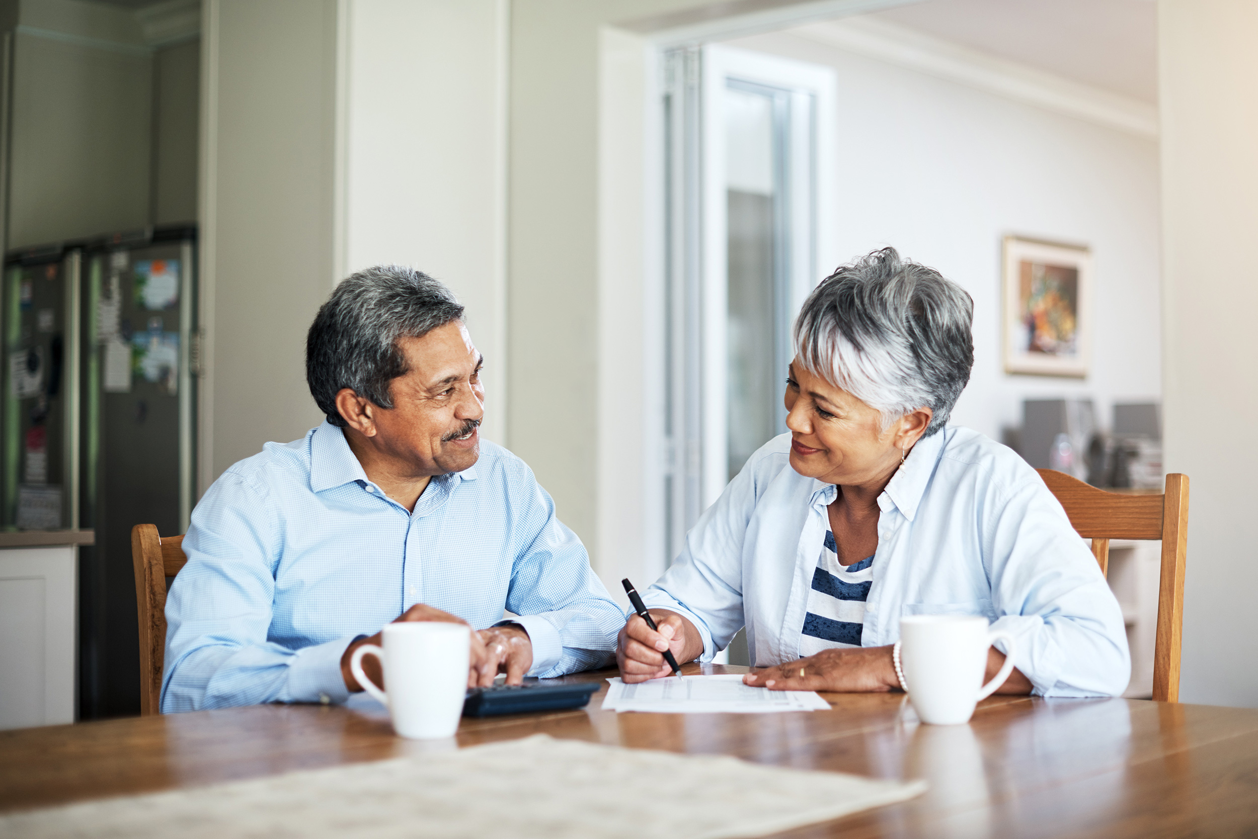 Sitting at a dining room table with papers, a calculator and coffee mugs, a man and woman smile at each other. Social Security Disability payments can change over time.