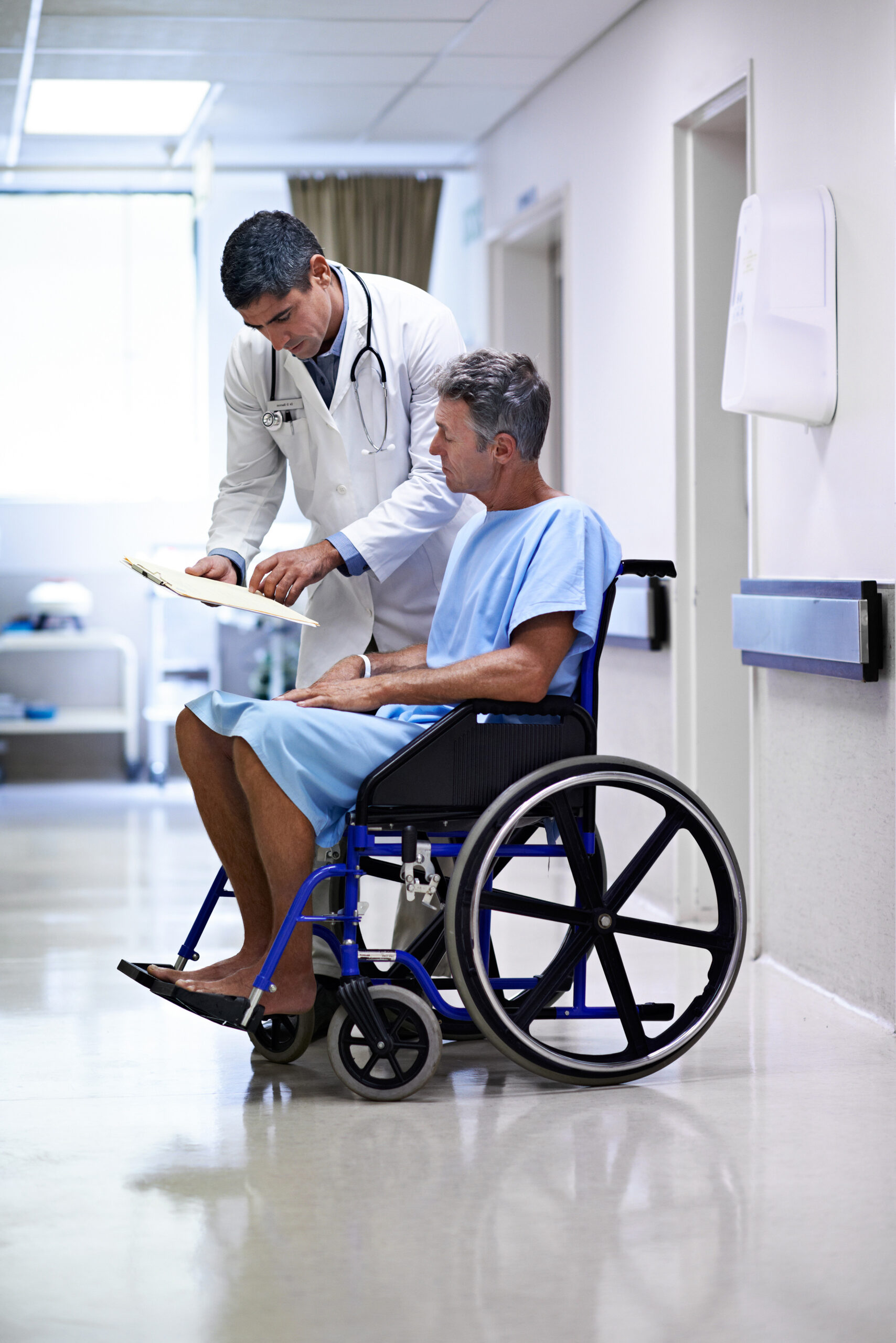 A man in a hospital gown sitting in a wheelchair listens to his doctor, who leans next to him with a clip board. If you have life-changing health problems, the Oklahoma disability lawyers at Troutman & Troutman can help you seek financial relief.