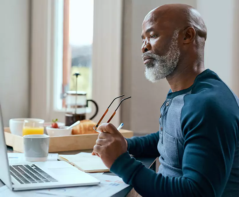 An older man taking off his glasses and looking at his computer screen.